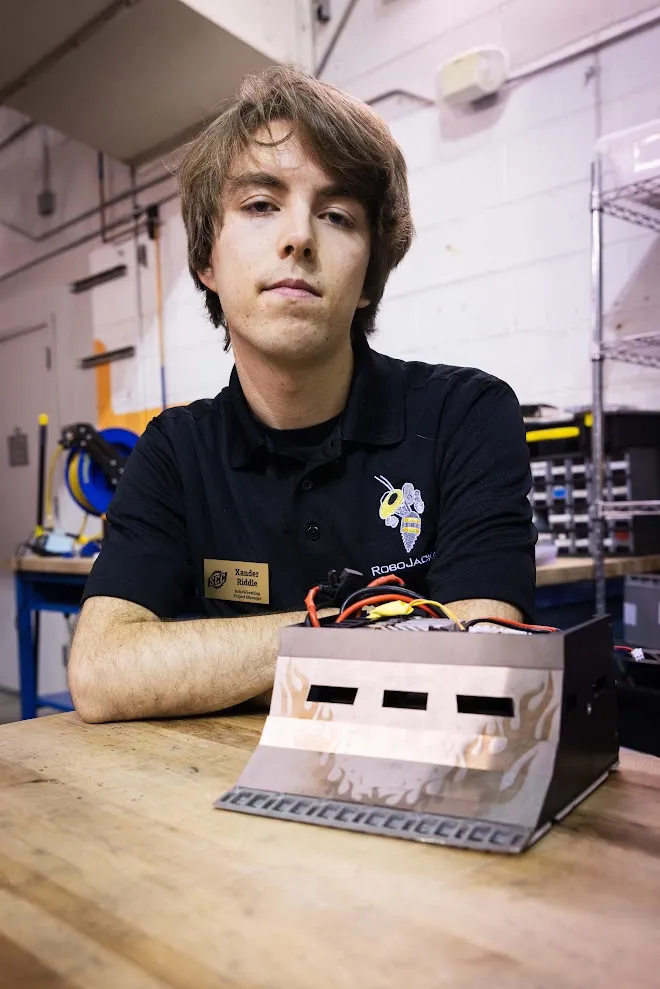 Xander in a RoboJackets polo at a workbench beside a flame-painted combat robot